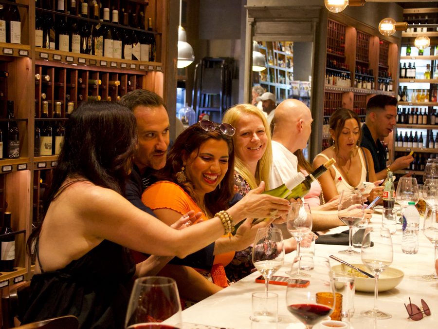 A group laughing over wine at a long table in a wine shop